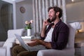 Young attractive smiling guy is browsing at his laptop and talking oh mobile phone, sitting at home on the cozy sofa, wearing Royalty Free Stock Photo