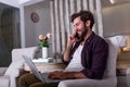 Young attractive smiling guy is browsing at his laptop and talking oh mobile phone, sitting at home on the cozy sofa, wearing Royalty Free Stock Photo