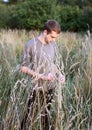 A young attractive thin guy standing in the tall grass on the forest background and looking down.