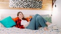 A young attractive white woman is lying on the bed and smiling in the modern bedroom while listening to musing using modern earbud