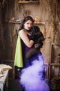 Young, attractive witch with a black cat in her hands is cooking a potion in a large black cauldron emitting magical purple smoke