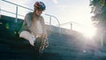 Young attractive woman adjusting rollerblades while sitting on the stairs in the skatepark. Direct sunshine