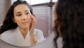 Young attractive woman in bathrobe looks in mirror in bathroom touches moisturized face after cosmetic procedure at home Royalty Free Stock Photo