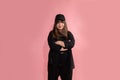 A young attractive woman in black baseball cap and sportswear stands on pink background in studio with her arms crossed over her