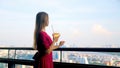 Young attractive woman drinking cocktail at luxury rooftop restaurant with high cityscape. Elegant female in red dress