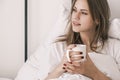 Young attractive woman lies in bed after waking up with white cup in her hand looking out the window. Morning routine Royalty Free Stock Photo