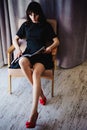 Young attractive woman with long legs in black elegant dress, sits in chair near window in interior of room