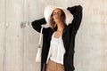 Young attractive woman in pretty fashionable beautiful clothes in trendy glasses with bag straightens straw summer hat near gray Royalty Free Stock Photo