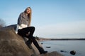 Young attractive woman sitting on a rock near the water. She`s wearing a gray sweater and black pants. She smiles. Blue sky Royalty Free Stock Photo