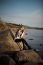 Young attractive woman sitting on a rock near the water. She`s wearing a gray sweater and black pants. She smiles. Blue sky Royalty Free Stock Photo