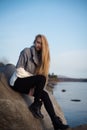 Young attractive woman sitting on a rock near the water. She`s wearing a gray sweater and black pants. She smiles. Blue sky Royalty Free Stock Photo