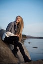 Young attractive woman sitting on a rock near the water. She`s wearing a gray sweater and black pants. She smiles. Blue sky Royalty Free Stock Photo