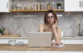 Young attractive woman sitting at the table and working on laptop in the kitchen at home Royalty Free Stock Photo