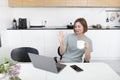 Young attractive woman sitting at the table and working and talking in a video call on laptop in the kitchen at home Royalty Free Stock Photo