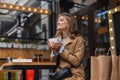 Young attractive woman in trench coat happily looking aside while holding white cup with tea in hand outdoor at cozy