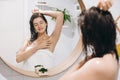 Young attractive woman with wet hair in white towel enjoying smooth skin after shaving armpits, reflection in mirror in stylish