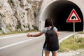 Young beautiful woman with black backpack hitchhiking standing near the road tunnel. Back view. Royalty Free Stock Photo