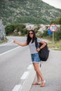 Young beautiful woman with black backpack hitchhiking standing on road. Beautiful young female hitchhiker by the road during vacat Royalty Free Stock Photo