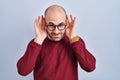 Young bald man with beard standing over white background wearing glasses trying to hear both hands on ear gesture, curious for Royalty Free Stock Photo
