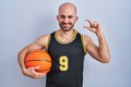 Young bald man with beard wearing basketball uniform holding ball smiling and confident gesturing with hand doing small size sign Royalty Free Stock Photo