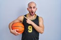 Young bald man with beard wearing basketball uniform holding ball surprised pointing with finger to the side, open mouth amazed Royalty Free Stock Photo