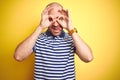 Young bald man with beard wearing casual striped blue t-shirt over yellow isolated background doing ok gesture like binoculars Royalty Free Stock Photo