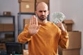 Young bald man with beard working at small business ecommerce holding money with open hand doing stop sign with serious and