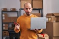 Young bald man with beard working at small business ecommerce with laptop in shock face, looking skeptical and sarcastic, Royalty Free Stock Photo