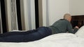 Young bald man in a gray sweater and jeans lies on a bed in a hotel room, includes a laptop, the concept of business travel,