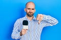 Young bald man holding reporter microphone with angry face, negative sign showing dislike with thumbs down, rejection concept Royalty Free Stock Photo