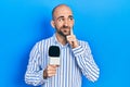 Young bald man holding reporter microphone serious face thinking about question with hand on chin, thoughtful about confusing idea Royalty Free Stock Photo