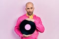 Young bald man holding vinyl disc in shock face, looking skeptical and sarcastic, surprised with open mouth