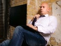 Young bald man wearing suit and tie using laptop while sitting near the damaged plaster wall. Businessman unbuttons his shirt and Royalty Free Stock Photo
