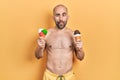 Young bald man wearing swimwear holding two ice cream cones making fish face with mouth and squinting eyes, crazy and comical