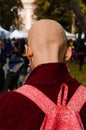 Young bald woman with long earrings. Dressed in a burgundy coat with a pink backpack. Back view. Outdoors