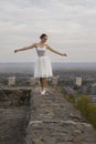 Young ballerina in white dress and satin ballet shoes posing on the edge of old fortress wall on a grey sky background Royalty Free Stock Photo