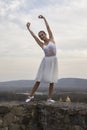 Young ballerina in white dress and satin ballet shoes posing on the edge of old fortress wall on a grey sky background Royalty Free Stock Photo