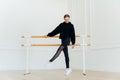 Young ballet dancer has warm up, stands near handrails, dressed in black sportclothes, stands on tip toe, wears white sneakers,