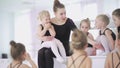 Young ballet teacher sitting on bench in ballet school with little girls in black leotards and communicating with them