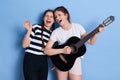 Young band rehearsing, girls in casual attires singing songs loudly, standing against blue wall, females having fun while playing