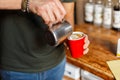 Young barista male making latte coffee in vintage coffee shop. Men`s hands are holding a metal cup and a paper red coffee cup. Royalty Free Stock Photo