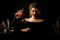 Young bartender woman in black blouse holds glass and pours cocktail into it.