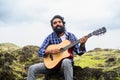 Young beard man playing guitar by singing song while sitting on top of hill - concept of hobbies, leisure activity and Royalty Free Stock Photo