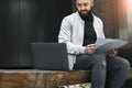 Young bearded business man working on laptop while sitting on bench outdoors. Man holds paper documents in his hands. Lifestyle Royalty Free Stock Photo