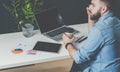 Young bearded businessman in denim shirt is sitting at office at table, talking on mobile phone and drinking coffee. Royalty Free Stock Photo