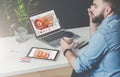 Young bearded businessman in denim shirt is sitting at office at table, talking on mobile phone and drinking coffee. Royalty Free Stock Photo