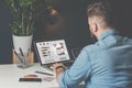 Young bearded businessman in denim shirt is sitting in office at table and is using laptop with charts, graphs