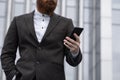 Young bearded Businessman holding mobile smartphone using app texting sms message wearing jacket outdoor. Successful Royalty Free Stock Photo