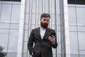 Young bearded Businessman holding mobile smartphone using app texting sms message wearing jacket outdoor. Successful Royalty Free Stock Photo