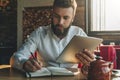 Young bearded businessman sits in cafe, home at table, holding tablet computer and writes in notebook. Royalty Free Stock Photo